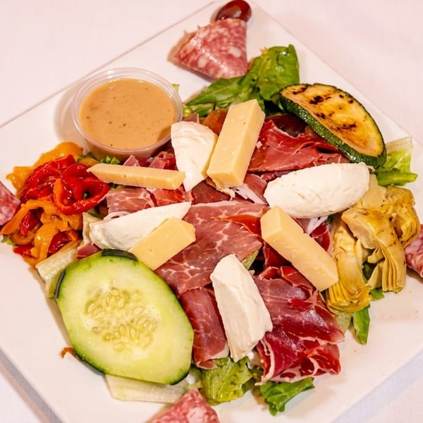 Antipasto salad with meats, cheeses, grilled vegetables, and dressing on a white plate.
