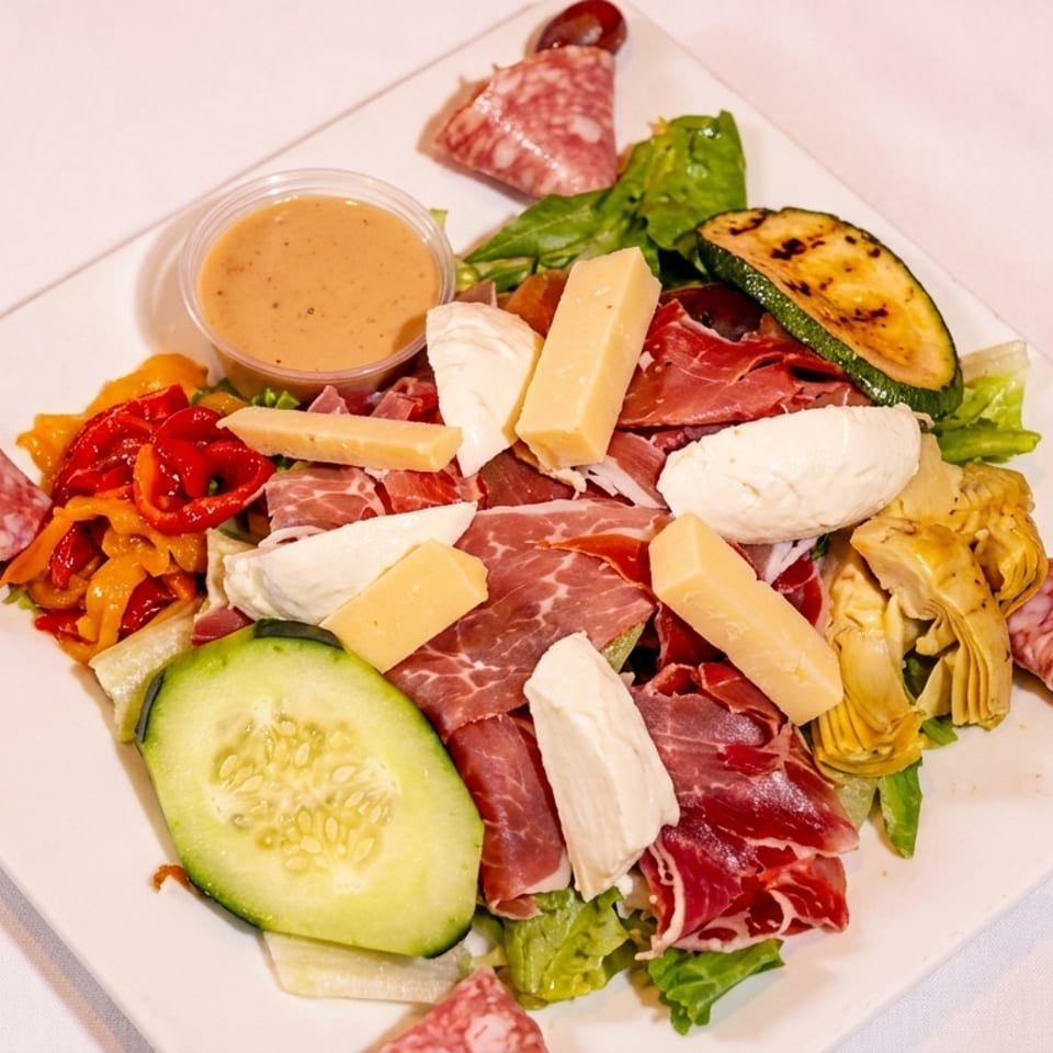 Antipasto salad with meats, cheeses, grilled vegetables, and dressing on a white plate.