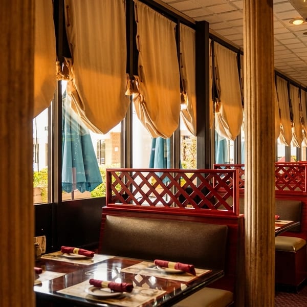 Restaurant interior with booths, red lattice dividers, large windows with cream drapes, and decorative columns.