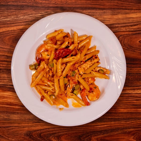 Penne pasta with shrimp, grilled chicken, vegetables, and sun-dried tomatoes in a creamy orange sauce on a white plate.