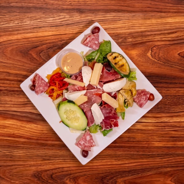Antipasto platter with assorted cured meats, cheeses, vegetables, olives, and dipping sauce on a white plate atop a wooden table.