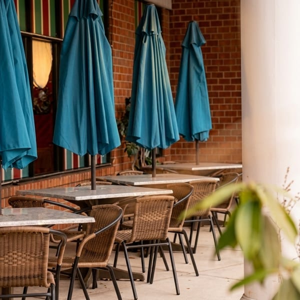 Outdoor patio with closed blue umbrellas, wicker chairs, and marble tables against a brick wall.