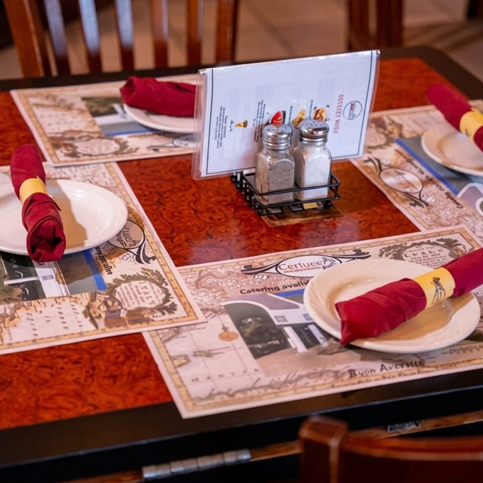 A restaurant table set with placemats, white plates, red napkins, and a menu with salt and pepper shakers.